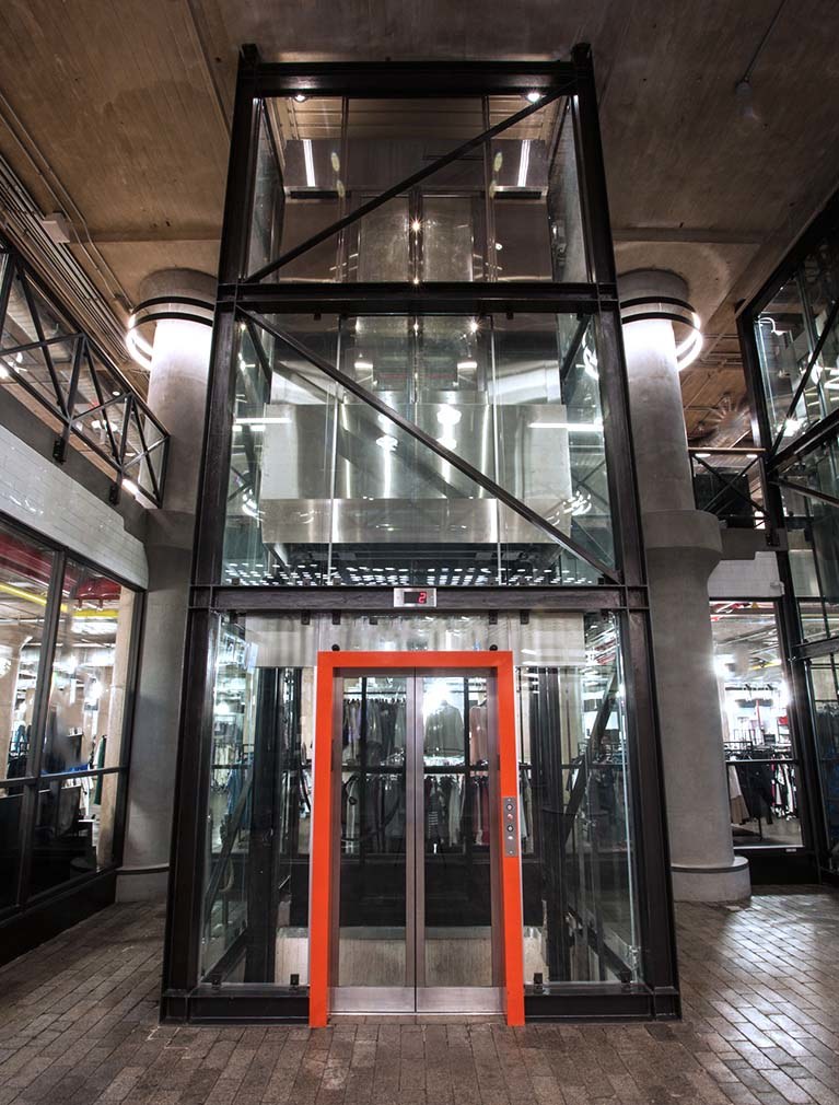 850 Third Ave, Brooklyn, straight on from lobby, glass elevator with black steel girders & orange accent door frame