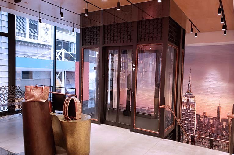 Coach custom glass elevator seamlessly integrates into the showroom with NYC skyline painting behind and handbag displays surrounding